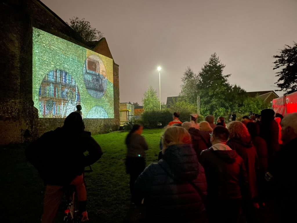 A photo of an outdoor projection, a brick wall of a building on the left is Everton Library and an illustrated image of books and a computer is being projected onto it. A crowd of people are gathered to watch the projections. It is late evening and in a park with grass and trees in the background.