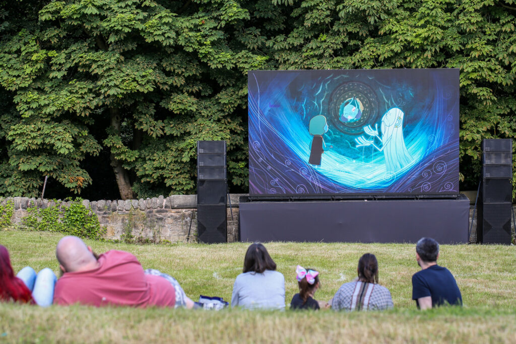 A photo of an outdoor cinema, a large LED screen showing the film Song of the Sea, with a group of people sitting on grass watching the screen. The image on the screen is of an animation, a small boy is talking is talking to a spirit looking into a swirl of blue water.
